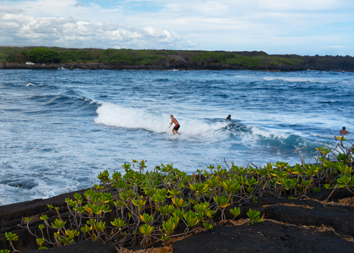 Black sand beach on Hawaii Island with a man surfing in the ocean.