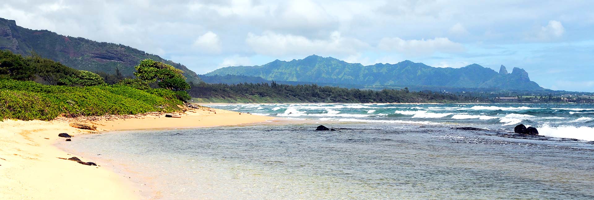 Beach shoreline off Kauai with green foliage on the shore and green mountains in the background
