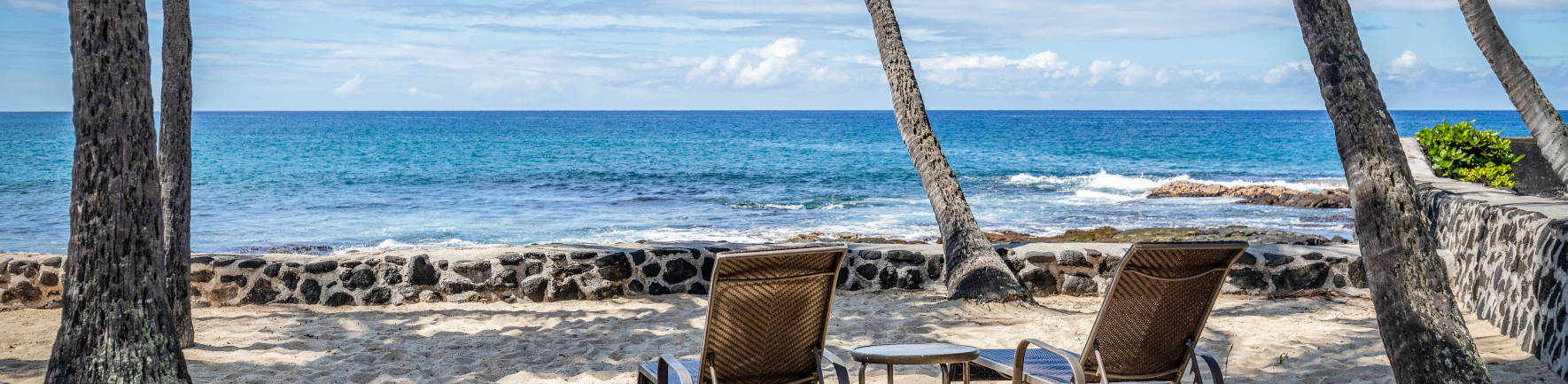 Two lounge chairs near the shore break facing the ocean under some coconut trees at the Aston Kona by the Sea