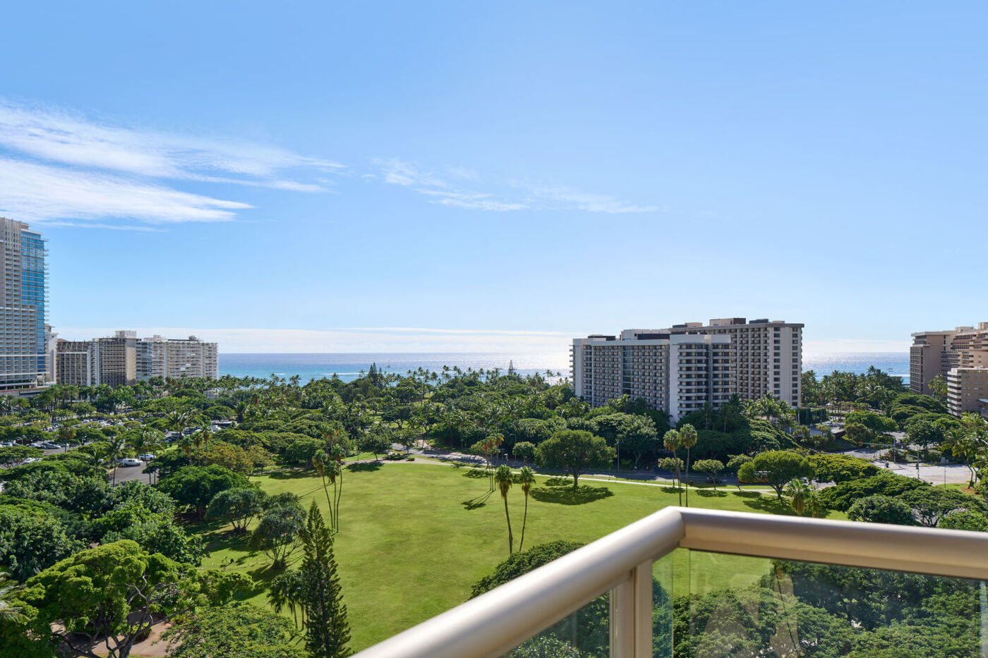 Balcony view of the park with green grass, and variety of trees, cityscape view, and pacific ocean in the horizon.