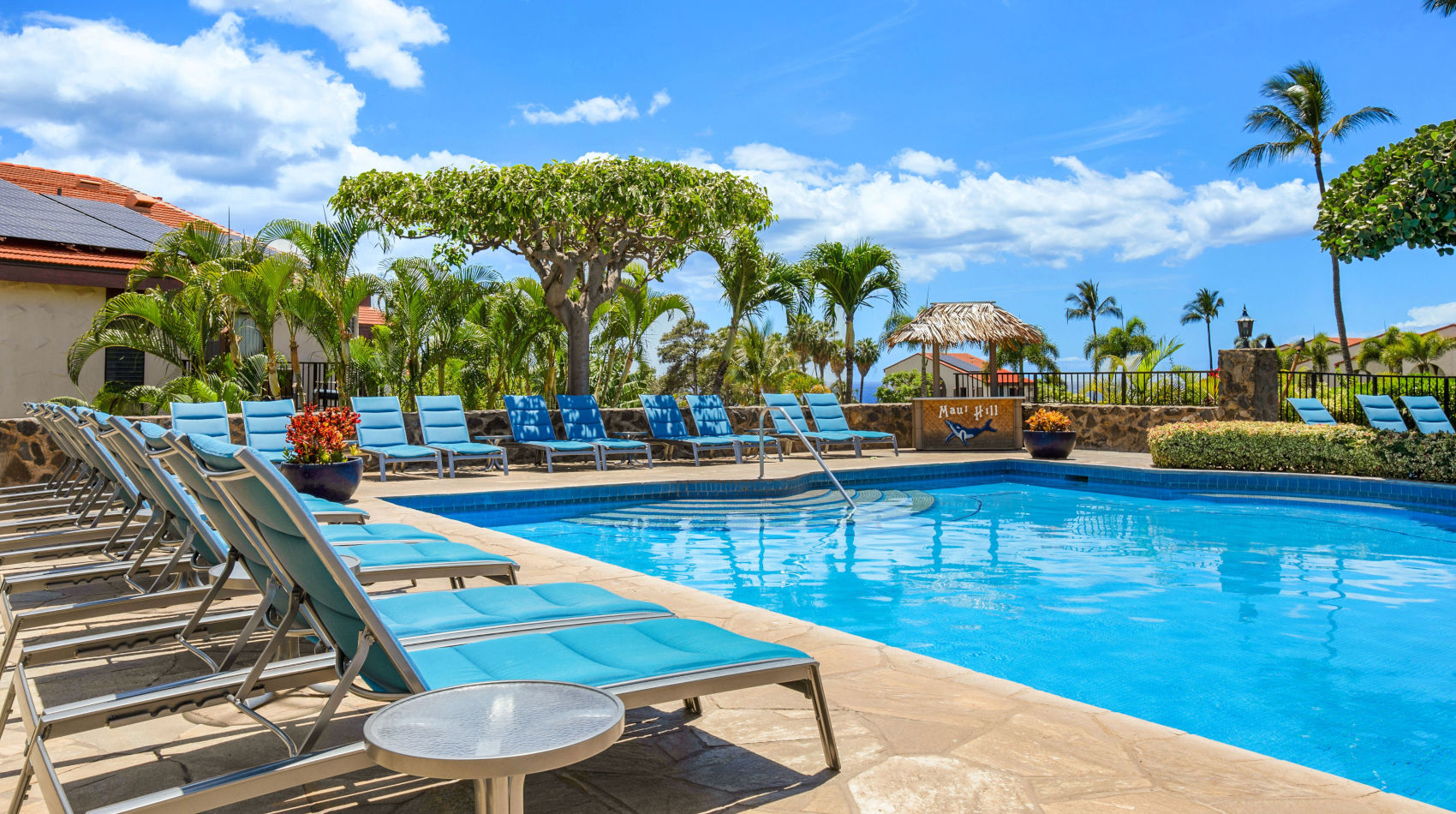 Pool at the Maui Hill with blue padded lounge chairs, landscaped with lush green palm trees and colorful bushes and accommodation building units in the background