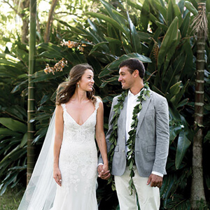 Kalii Scheer wearing a white wedding gown holding hands with her husband wearing a grey jacket over a white shirt and pants wearing a maile lei